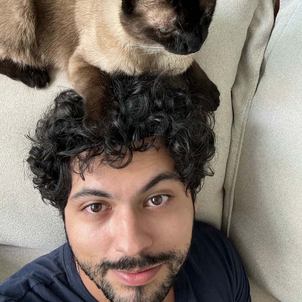 Ian lying on a couch, looking up at the camera, with his Siamese cat perched on top of his head.